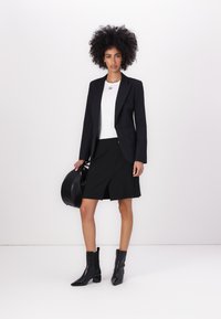 Černý zakázkový blazer na bílé upnuté halence, doplněný černou zavinovací sukní. Model má na sobě černé kotníkové boty a drží kulatou kabelku.