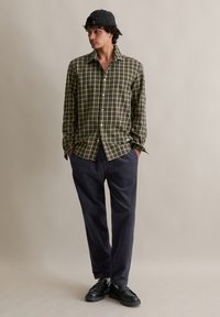 Jongeman in een groen geruit shirt, marineblauwe corduroybroek, zwarte schoenen en een grijze aangekapte pet, staand tegen een effen achtergrond.