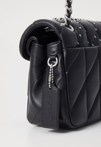Czarna pikowana skórzana torebka na ramię z zaokrąglonym dnem, srebrnym łańcuszkiem i wyraźną metką. Zawiera małe ozdobne ćwieki wzdłuż szwów.