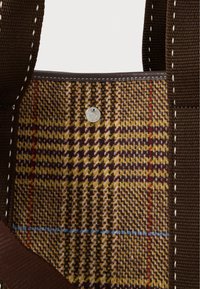 Κοντινή λήψη μιας καφέ τσάντας από καρό ύφασμα με υφασμένη υφή, καφέ ιμάντες και ένα μικρό στρογγυλό μεταλλικό κουμπί χαραγμένο με κείμενο.