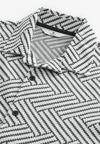 Camisa polo en blanco y negro con un patrón texturizado de espiga, que cuenta con un cuello doblado, tres botones negros y una etiqueta en el interior del cuello.