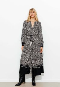 Robe à motif paisley noir et blanc avec une taille à nouer et des manches longues. Elle présente un ourlet noir avec une broderie décorative. Portée avec des bottes à talons noires.