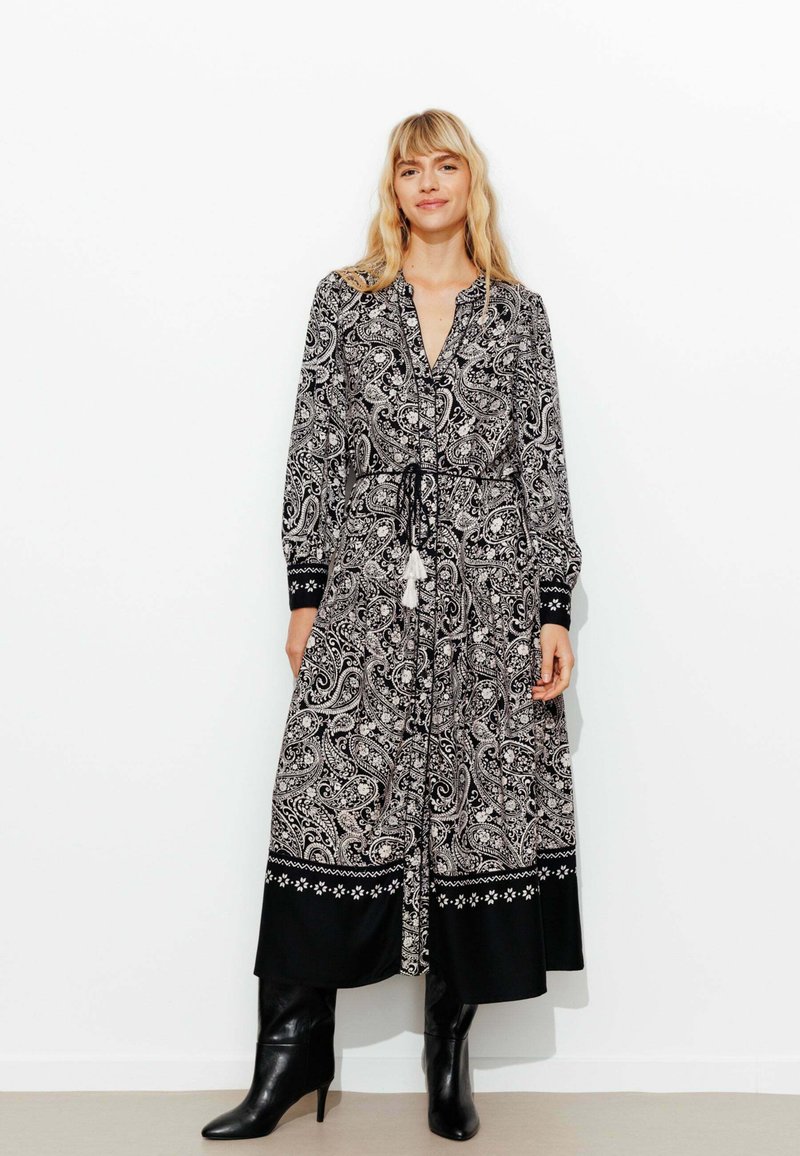 Robe à motif paisley noir et blanc avec une taille à nouer et des manches longues. Elle présente un ourlet noir avec une broderie décorative. Portée avec des bottes à talons noires.