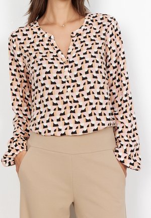 Kobieta ubrana w bluzkę z długim rękawem o geometrycznym wzorze w kolorach czarnym, różowym, białym i beżowym, zestawioną z beżowymi spodniami z wysokim stanem.