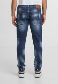 Modré džíny z denimu se zesvětleným povrchem, mírným opotřebením na zadních kapsách a hladkou strukturou. Obsahují hnědý kožený štítek na pase.