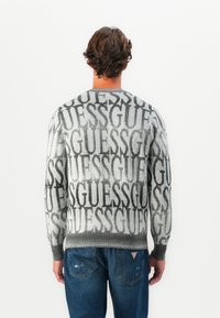 Uomo con capelli castani mossi che indossa un maglione grigio con testo ripetuto "GUESS" e jeans blu, in piedi di fronte a un muro bianco.