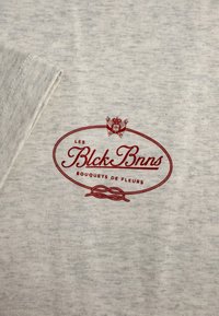 Lichtgrijze t-shirt met een zachte textuur, voorzien van een rood ovaal logo met de tekst "LES Blck Bnns Bouquets de Fleurs" en decoratieve accenten.