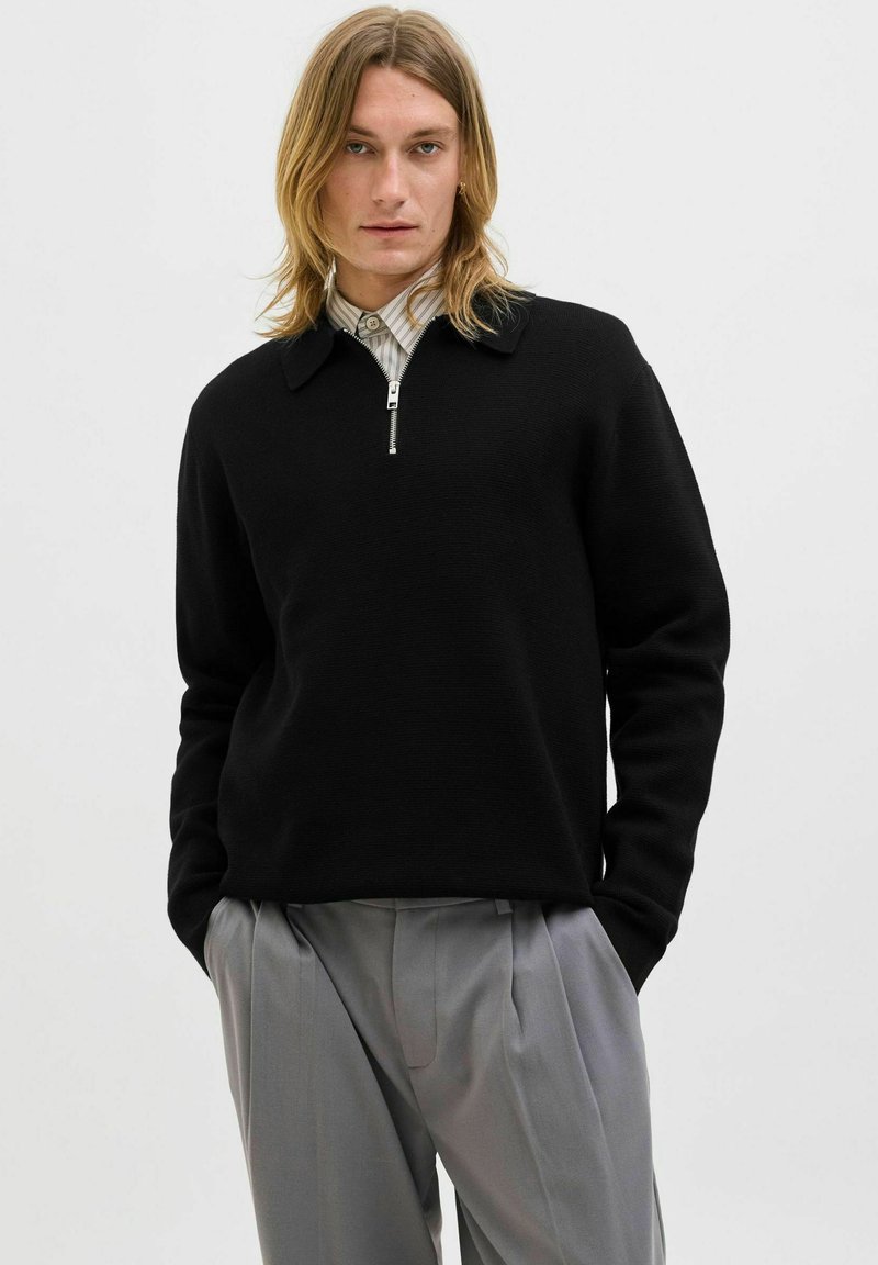 Mladý člověk s dlouhými vlasy, oblečený v černé zipové mikině a šedých skladaných kalhotách, s rukama v kapsách, na neutrálním pozadí.