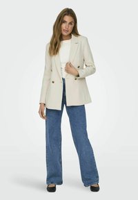 Blazer doppiopetto beige chiaro con bottoni dorati, spalle strutturate, abbinato a jeans blu a gamba larga e scarpe nere. Top bianco sotto.