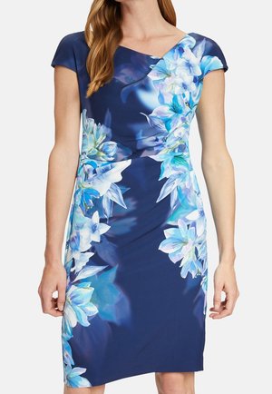 Kvinde iført en tætsiddende mørkeblå kjole med kasketærmer og store lyseblå og hvide blomsterprint langs siden og skulderen.