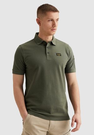 Junger Mann trägt ein olivgrünes Poloshirt mit einem kleinen schwarzen und gelben Aufnäher auf der Brust, kombiniert mit beigen Hosen.
