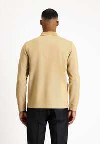 Beige Langarmhemd mit strukturiertem Strickdesign, Kragen, und gerippten Manschetten, kombiniert mit schwarzen Hosen.