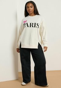 Sudadera crema con "PARIS" impreso en negro y un corazón rosa. Presenta un corte holgado, aberturas laterales y puños acanalados. Combinada con pantalones vaqueros de pierna ancha en negro.