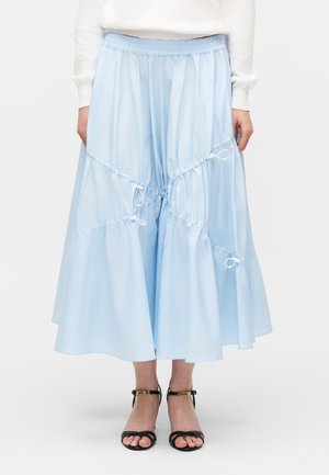 Svetlomodrá midi sukňa so zhromaždenými, nariasenými detailmi a šnúrkami na zaväzovanie, nosená s bielym topom a čiernymi sandálmi na podpätku s otvorenou špičkou a remienkom okolo členka.