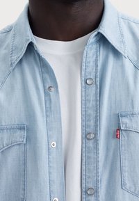 Světle modrá džínsová košile se špetlovými knoflíky, krátkým límečkem a dvěma náprsními kapsami. Pod ní je vidět bílý tričko s kulatým výstřihem.