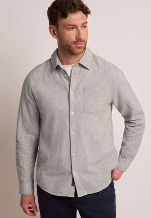 Man die een lichtgrijs overhemd met lange mouwen en knopen draagt over een wit onderhemd, met een donkere broek, kijkt naar links met één hand in zijn zak.