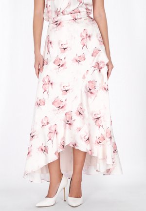 Frau trägt einen weißen High-Low-Rock mit pinken Blumen und weißen High-Heels vor einfachem Hintergrund.