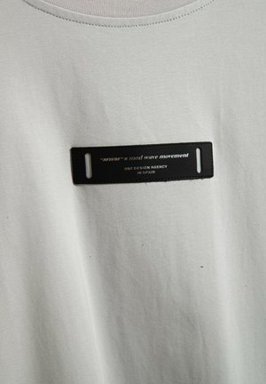 Camiseta de algodón blanca con una etiqueta rectangular negra con texto plateado que dice "mod wave movement" y "OBF DESIGN AGENCY EN ESPAÑA."