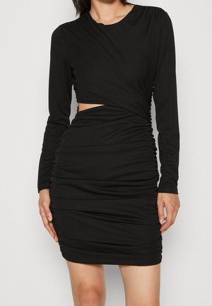 Femme portant une robe noire moulante à manches longues avec une découpe latérale et des détails froncés sur la jupe et le haut, sur fond blanc.