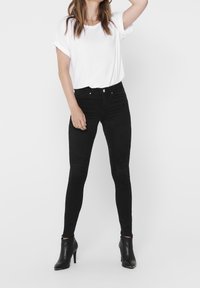 Donna che indossa una t-shirt bianca a maniche corte infilata in jeans skinny neri a vita alta e stivaletti neri con tacco, posando con un braccio alzato.