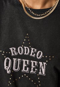 Camiseta de algodón negra con gráfico "RODEO QUEEN" en fuente blanca desgastada, acentuada con tachuelas doradas redondas dispuestas en un patrón de estrella.