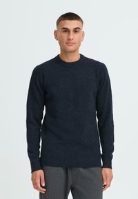 Marineblauer Strickpullover mit langen Ärmeln, Rundhalsausschnitt und geripptem Saum. Weiche Textur und lässige Passform. Getragen mit grauen Hosen.