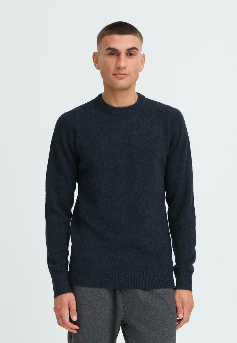 Marineblauer Strickpullover mit langen Ärmeln, Rundhalsausschnitt und geripptem Saum. Weiche Textur und lässige Passform. Getragen mit grauen Hosen.