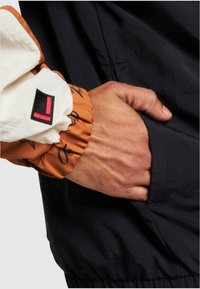 Farverig jakke med en sort krop og orange mønstrede ærmer. Har en elastisk manchet og et logo-patch på hvidt stof. Hånd i lommen.