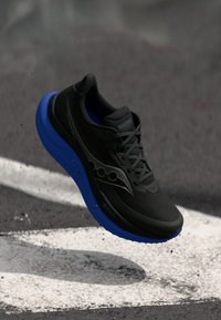 Zwarte en blauwe hardloopschoen met een gestructureerde zool die boven het asfalt zweeft met witte wegmarkeringen.