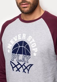 Chemise grise à manches longues avec des manches raglan bordeaux. Présente le texte "NEVER STOP PLAY" et un graphique de basketball en bleu marine. Tissu doux au toucher.