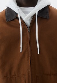 Brązowa kurtka zapinana na zamek z ciemnym kołnierzem, noszona na jasnoszarym hoodie. Posiada białe sznurki i duże kieszenie z przodu. Gładka struktura materiału.