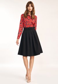 Kobieta w czerwonej kwiatowej bluzce z długim rękawem schowanej w czarną spódnicę plisowaną o wysokim stanie z paskiem oraz w cielistych szpilkach.