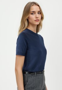 Marineblaues kurzärmeliges Strickoberteil mit Rundhalsausschnitt, glatter Textur und figurbetonter Passform, kombiniert mit grauen Nadelstreifen-Hosen und einem schwarzen Gürtel.