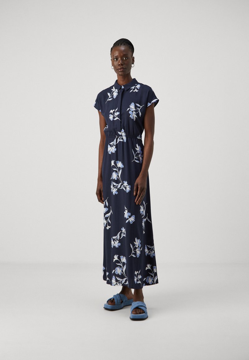 Abito maxi blu navy con maniche corte a cappuccio, stampa floreale in azzurro chiaro e bianco, vita arricciata, indossato con sandali blu chiaro.