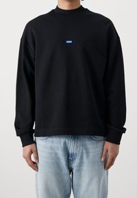Persona que lleva una sudadera negra de manga larga con un pequeño logo azul "HUGO" en el pecho, combinada con jeans de mezclilla azul claro.