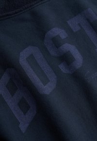 Tissu bleu marine avec des lettres en blocs bleu foncé formant le mot "BOST" visible, montrant la texture et de légers plis du tissu.