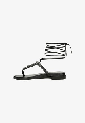 Zwarte platte sandalen met een enkelriem en decoratieve metalen accenten. Beschikt over een slingerende vetersluiting rond de enkel. Glad textuur.