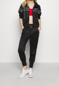 Zwarte sportieve trainingsset met een zip-up jas met witte logodetails en zwarte broek met witte zijkanten; gedragen met witte sneakers.