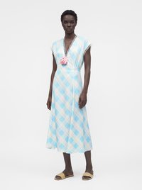 Γυναίκα φοράει μπλε και λευκό καρό midi φόρεμα με δαντέλα και μια ροζ υφασμάτινη καρφίτσα σε σχήμα λουλουδιού, συνδυασμένο με μπεζ σανδάλια.