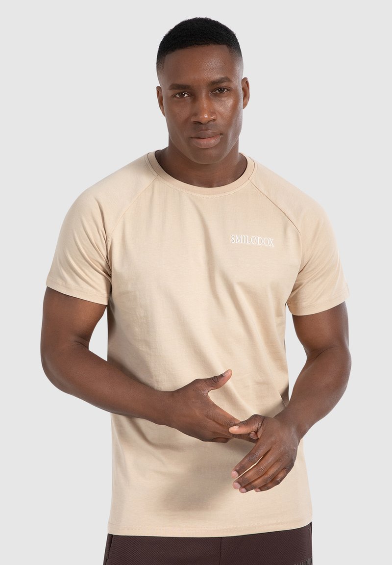 Beżowy t-shirt z krótkim rękawem, okrągłym dekoltem i rękawami raglanowymi. Posiada subtelne białe logo "SMILODOX" na piersi. Gładka tekstura materiału.