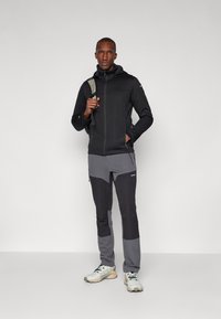 Giacca nera con zip e cappuccio, pantaloni grigi e neri con toppe sulle ginocchia, abbinati a scarpe da ginnastica di colore chiaro.
