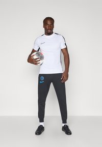 White short-sleeve Nike shirt with black accents, grey sweatpants featuring a blue logo, holding a patterned soccer ball, with black shoes.