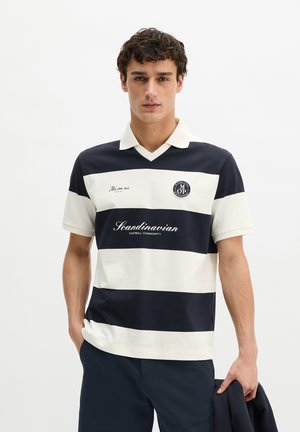 Jeune homme portant un polo à manches courtes rayé bleu marine et blanc avec le texte « Scandinavian Football Community », tenant une veste bleu marine dans sa main droite.