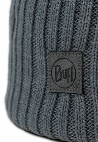 Gorro de lana gris con ribetes verticales. Presenta un parche de tela gris oscuro con el logo "Buff". Textura suave y diseño ajustado.