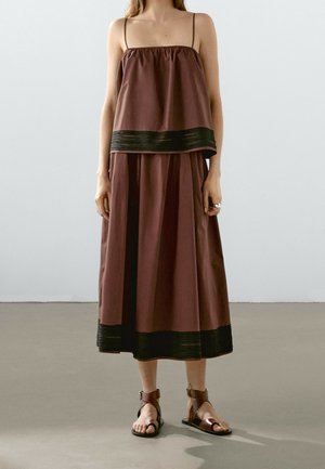 Femme portant une robe marron sans manches avec un bord noir à la taille et à l’ourlet, associée à des sandales plates marron, debout sur un sol gris.