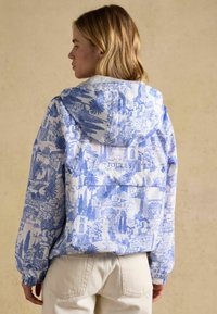 Naine kannab valge ja sinise mustriga kapuutsiga jope, millel on maastikupildid, seisab selili jaotunud beežilt ühtlasel taustal.