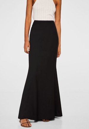 Femme portant une jupe longue noire taille haute évasée avec un top blanc sans manches côtelé et des sandales noires à lanières.