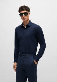 Navy long-sleeve button-up shirt in smooth fabric, paired with matching tailored trousers and dark sunglasses, featuring a modern cut.