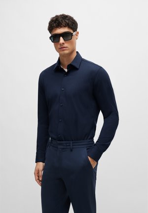 Camisa de botones de manga larga en color navy, fabricada en un tejido suave, combinada con pantalones a medida a juego y gafas de sol oscuras, presentando un corte moderno.