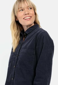 Granatowa koszula z korduroju z zapięciem na guziki, dwoma kieszeniami na piersi i teksturowanym materiałem; posiada czarne guziki i luźny krój.